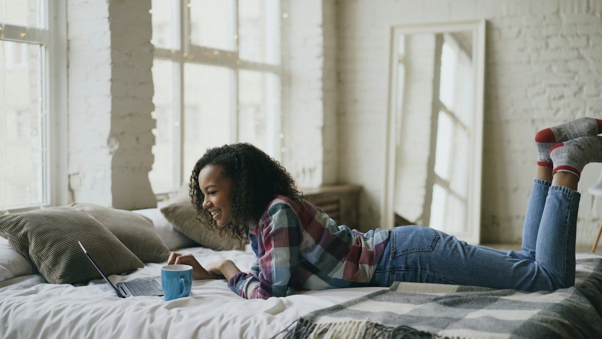 Curly mixed race student girl lauging while using laptop for sharing social media lying in bed at home